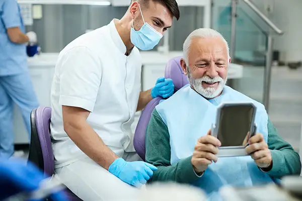 Happy, senior, white patient looking at himself in a mirror with a pleased, white male dental assistant Happy, senior, white patient looking at himself in a mirror with a pleased, white male dental assistant