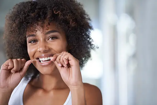 Young woman smiling while flossing her teeth, promoting good oral hygiene practices in a bright setting.